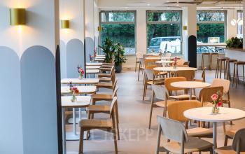 Een lichte zithoek met houten tafels en stoelen, ingericht als een lounge, geschikt voor informeel overleg of pauzes in een kantoorpand aan de Apollolaan 151 in Amsterdam Oud Zuid.