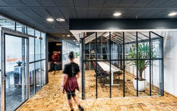 Modern kantoorpand gelegen op Johan Huizingalaan 400, Amsterdam Riekerpolder, met glazen vergaderruimte en houten vloer. Een persoon loopt door de ruimte. Ideaal voor bedrijven die kantoorruimte willen huren.