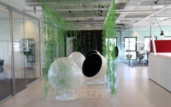 Moderne kantoorruimte huren op John M Keynesplein 12-46, Amsterdam Riekerpolder, met glazen wanden, bijzondere witte stoelen en hangende groene decoratie. Medewerkers werken aan bureaus.