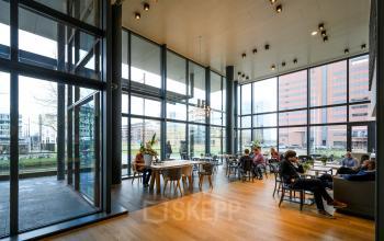 Modern ingerichte kantoorruimte te huur op Thomas R. Malthusstraat 1-3 in Amsterdam Riekerpolder met grote ramen, houten vloeren, en mensen die vergaderen en werken in een ruime, lichte omgeving.