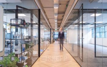 Modern kantoorpand aan de Teleportboulevard 120 in Amsterdam Sloterdijk met glazen wanden en een houten vloer, waar iemand door de gang loopt. Ideaal voor kantoorruimte huren.