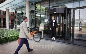 Twee personen voor het kantoorpand op Teleportboulevard 110 in Amsterdam Sloterdijk, een persoon komt naar buiten terwijl een ander met een fiets richting de ingang loopt.