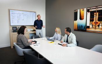 Zakelijke bijeenkomst in een moderne vergaderruimte op Teleportboulevard 110, Amsterdam Sloterdijk. Vier personen, waaronder een presentator, bespreken werkdocumenten bij een lange tafel met laptops en scherm. Kantoor huren mogelijk.