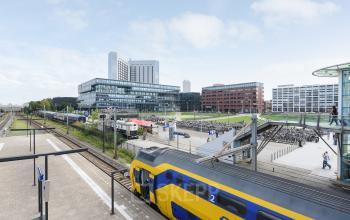 Buitenzijde van kantoorpand op Teleportboulevard 110 in Amsterdam Sloterdijk, met nabijgelegen treinsporen en een gele NS-trein. Ideale locatie voor kantoorruimte huren met uitstekende openbaar vervoer verbindingen.