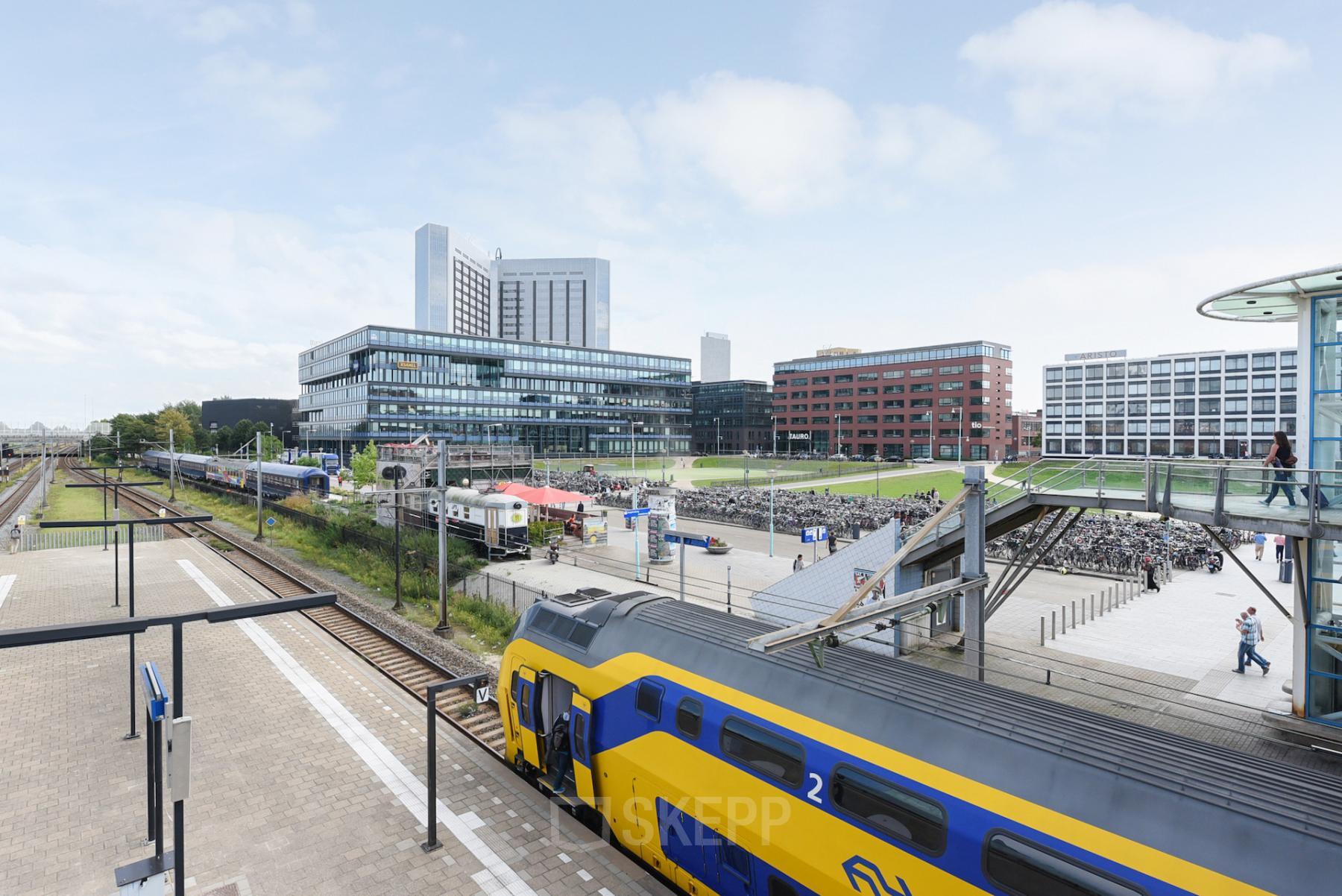 Moderne kantoorpanden gezien vanaf Teleportboulevard 110 in Amsterdam Sloterdijk, met een levendig stationsplein en voorbijrijdende treinen.