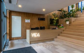 Ingang van een kantoorpand op Kabelweg 57, Amsterdam Sloterdijk. Moderne receptie met houten balie en trappen, omgeven door planten. Kantoorruimte huren met een uitnodigende uitstraling.