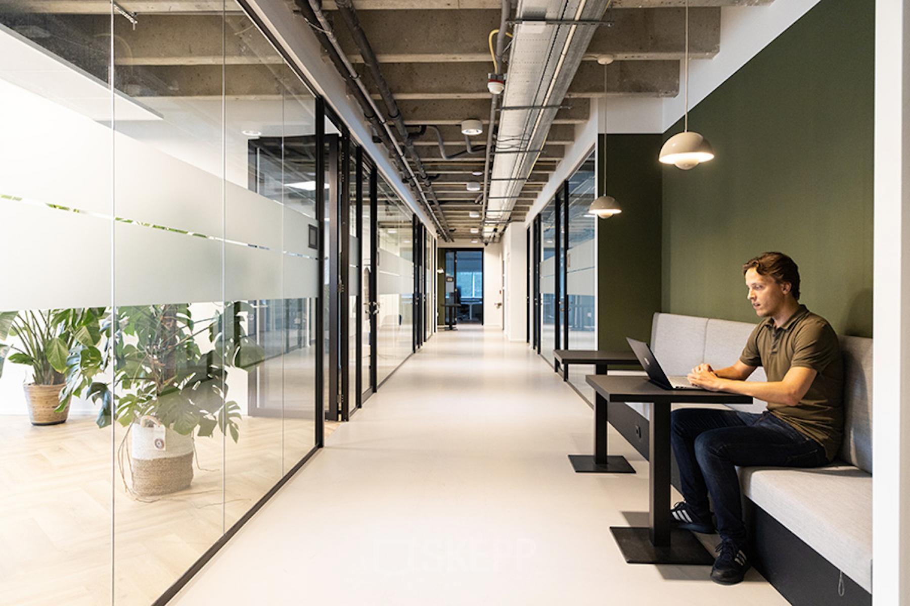 Modern kantoorpand op Kabelweg 37 in Amsterdam Sloterdijk met glazen wanden en loungegedeelte. Persoon werkt op laptop aan tafel. Ideaal voor kantoorruimte huren.