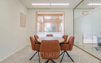 Vergaderruimte met zes oranje stoelen rond een ronde tafel, grote ramen bieden veel natuurlijk licht in een kantoorpand op Basisweg 32, Amsterdam Sloterdijk, ideaal voor verhuur.