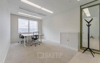 Lichte kantoorruimte op Basisweg 32, Amsterdam Sloterdijk, met twee bureaus en stoelen, ideaal om kantoor te huren. Minimalistisch ingericht met neutrale tinten en een groot raam voor natuurlijk licht.