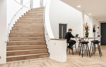 Moderne kantoorruimte op Arlandaweg 92 in Amsterdam Sloterdijk met houten trap, hoge tafels en zitkrukken. Een persoon werkt aan een laptop aan een hoge tafel met bloemenvaas.