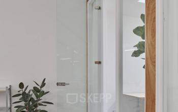 Kantoorruimte huren op Hondiusstraat 7H, Amsterdam West. Heldere ruimte met glazen deur, houten accenten en planten, ideaal voor inspirerende werkzaamheden in een moderne omgeving.