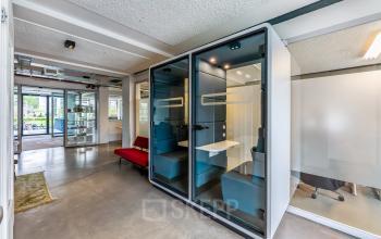 Moderne kantoorruimte te huur in Amsterdam West met glazen cabine voor privégesprekken en een rode bank in een open vloerplan. Het kantoorpand biedt gemakkelijke toegang tot faciliteiten. Ideaal voor flexwerken.