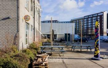 Buitenzijde van kantoorpand Rigakade 10 in Amsterdam West. Beton, tafels, en een kleurrijk standbeeld op de voorgrond. Geschikt voor kantoorruimte huren.