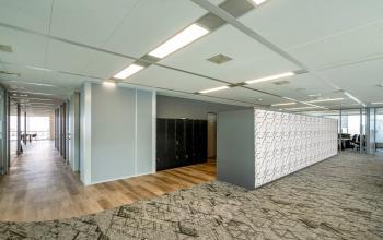 Binnenkant van kantoorpand gelegen op De Entree 230-266, Amsterdam Zuid-Oost. Ruime en moderne werkomgeving met lichtgrijze wanden, lockers en zachte vloerbedekking. Goed verlichte paden verbinden verschillende kantoorsecties.