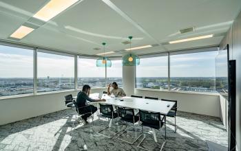 Vergaderruimte in kantoorpand op De Entree 230-266 Amsterdam Zuid-Oost met grote ramen, moderne verlichting en uitzicht over de stad. Twee personen vergaderen aan een rechthoekige tafel.