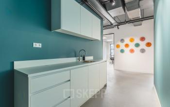 Moderne pantry in het kantoorpand op Paasheuvelweg 20, Amsterdam Zuid-Oost, met witte kasten en een blauwgroene muur. Voorzijde met wasbak en wanddecoratie in diverse kleuren. Ideaal voor kantoorruimte huren.