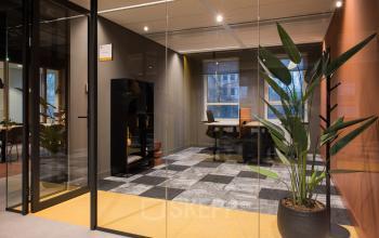 Moderne kantoorruimte huren in Amsterdam Zuid-Oost met glazen wanden en grijze vloertegels. Voorzien van een bureau, stoel en grote kamerplant, geschikt voor professionele activiteiten.