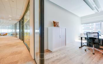 Moderne kantoorruimte te huur met glazen wanden en houten vloer, gelegen in Amsterdam Zuid-Oost. Het kantoorpand biedt uitzicht op de stad, met enkele bureaus en stoelen aanwezig.