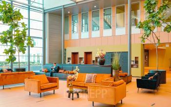 Kantoorruimte huren aan Jachthavenweg 109-A in Amsterdam Zuidas, met moderne zitbanken, een grote glazen gevel en groenvoorzieningen. Een persoon is bezig met een laptop.