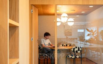 Moderne kantoorruimte huren in Amsterdam Zuidas met houten elementen en glazen wanden. Persoon werkt geconcentreerd op laptop in vergaderkamer. Gezellig en uitnodigend kantoorpand op Barbara Strozzilaan 101, ideaal voor zakelijke bijeenkomsten.