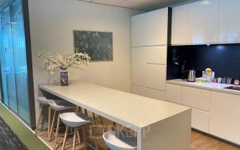 Moderne pantry in kantoorpand aan Claude Debussylaan 10, Amsterdam Zuidas, met witte hoogglans kasten, lange tafel met barkrukken en wanddecoratie.