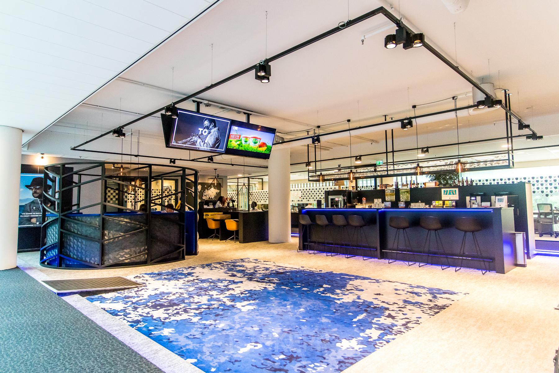 Moderne kantoorruimte te huur op Gustav Mahlerplein in Amsterdam Zuidas, met bar, kleurrijk tapijt en gestoffeerde zithoek.