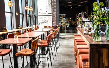 Moderne, lichte lunchruimte in een kantoorpand met hoge ramen en kleurrijke inrichting. Twee mensen zijn bezig met voorbereidende werkzaamheden aan de bar bij kantoorruimte huren in Apeldoorn.