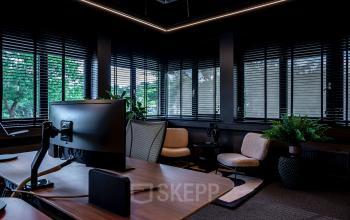 Donkere kantoorruimte met houten bureau en moderne stoelen, groene planten en grote ramen. Geschikt voor kantoorruimte huren.