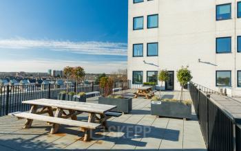 Dakterras van kantoorpand op Prins Willem Alexanderlaan 701, Apeldoorn met houten picknicktafels en plantenbakken. Ideaal voor een korte pauze tijdens het kantoorwerk. Helderblauwe lucht en omliggende stedelijke omgeving.