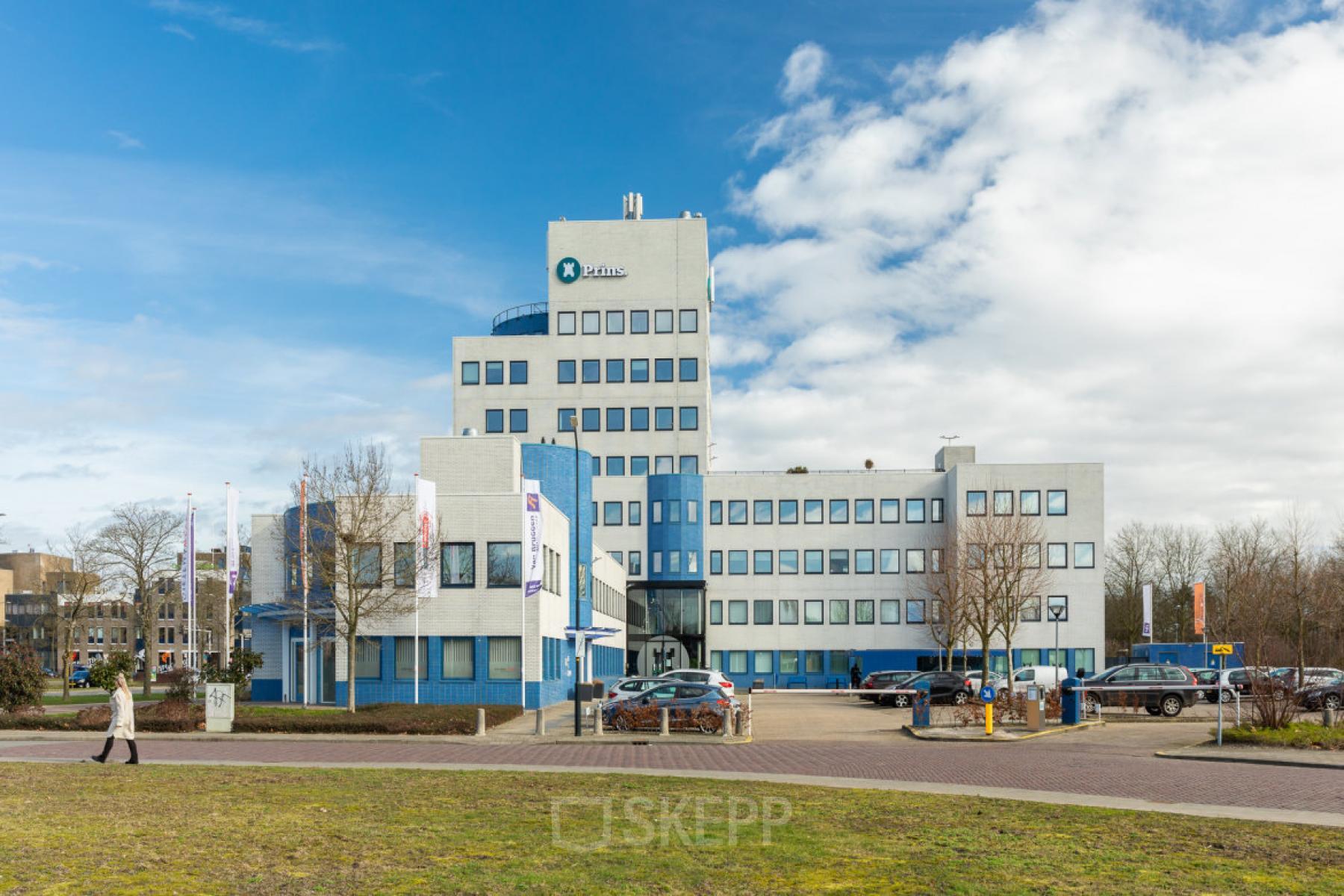 Exterieur van kantoorpand op Prins Willem Alexanderlaan 701, Apeldoorn. Ruime parkeerplaats voor het gebouw, waar mensen hun auto's stallen voor zakelijke bezoeken. Geschikte locatie voor kantoorruimte huren.