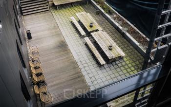 Terras van kantoorpand Roggestraat 111 in Apeldoorn met houten tafels en banken, rieten stoelen en plantenborders.