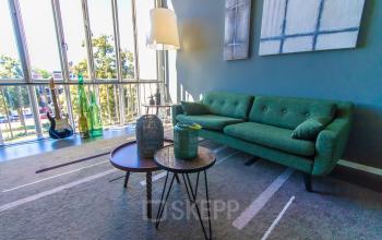 Stijlvolle kantoorlounge in Arnhem Centrum, met een groene bank, bijzettafels en grote ramen, voor huur op Jansbuitensingel 7.