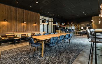 Arnhem IJsseloord Delta 1 kantoorruimte huren in een modern ingerichte kantine met lange houten tafel en blauwe stoelen, omringd door stijlvolle verlichting en industriële wandafwerking.