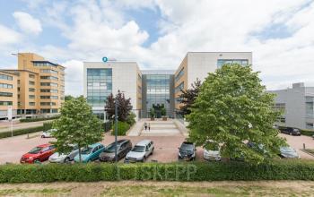 Vooraanzicht van het kantoorpand Delta 1, Arnhem IJsseloord met een parkeerterrein en grote ramen. Mensen lopen naar de ingang. Perfecte locatie om kantoorruimte te huren in een moderne omgeving.
