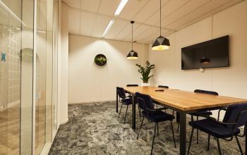 Moderne vergaderruimte in kantoorpand op Delta 1, Arnhem IJsseloord, met houten tafel, blauwe stoelen, hanglampen en muurtelevisie. Kantoorruimte huren voor efficiënte vergaderingen in een verfijnde sfeer.
