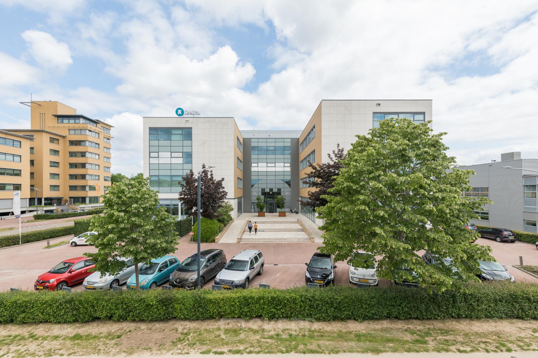Vooraanzicht van het kantoorpand Delta 1, Arnhem IJsseloord met een parkeerterrein en grote ramen. Mensen lopen naar de ingang. Perfecte locatie om kantoorruimte te huren in een moderne omgeving.