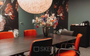 Stijlvolle kantoorruimte huren in Arnhem IJsseloord aan Vlamoven 34 met moderne vergadertafel, felgekleurde stoelen en decoratieve bloemenvaas.