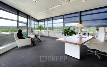 Zakelijke kantoorruimte huren op Mr D.U. Stikkerstraat 10, Arnhem Zuid; licht en ruim met grote ramen, ingericht met bureaus, stoelen, en planten. Twee mensen voeren een gesprek in de zithoek.