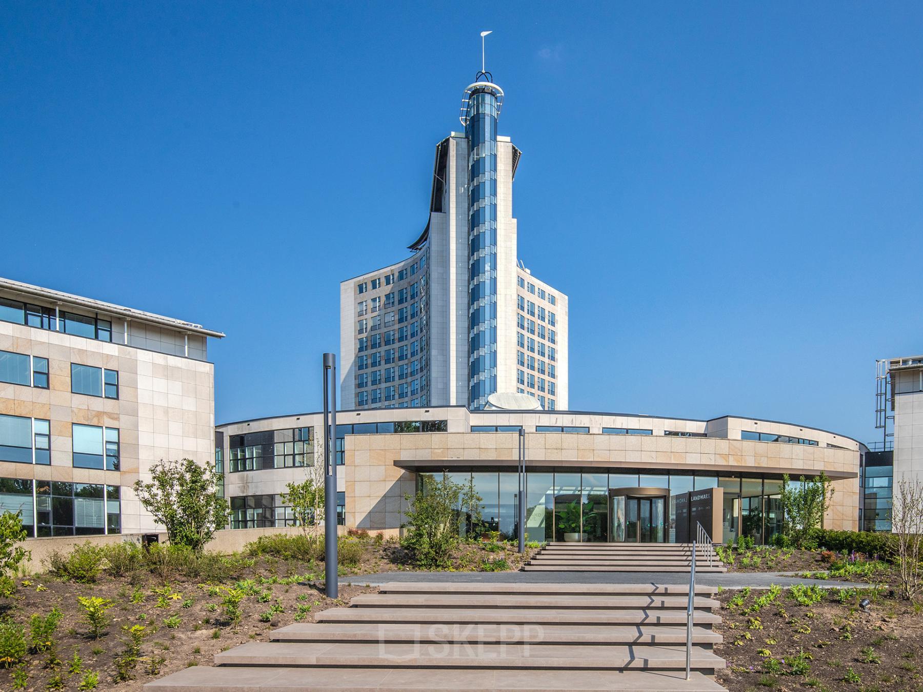 Exterieur van het kantoorpand gelegen op Rijksweg-West 2, Arnhem Zuid. Modern design met glazen gevels en een centraal hoog gebouw. Voorzijde met trappen en groenvoorziening.