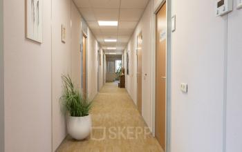 Lange gang in een kantoorpand met gesloten deuren aan beide zijden en een kantoorplant in een witte pot. Neutrale kleuren en tapijtbedekking. Locatie: Kamerlingh Onnesweg 10, Barendrecht.