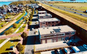 Luchtfoto van kantoor op Deltazijde 20b, Blaricum, met moderne bedrijfsgebouwen, groene omgeving en nabijgelegen snelweg. Perfecte locatie voor wie een kantoorruimte wil huren.