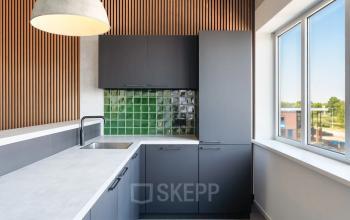 Moderne keuken in kantoorpand op Tolnasingel 1-3, Bodegraven, met grijskleurige kasten en groen betegelde achterwand, verlichting en groot raam. Geschikt voor korte pauzes in een open floorplan kantoor.