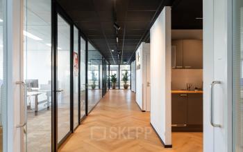 Verlichte gang in kantoorpand Tolnasingel 1-3, Bodegraven, met glazen wanden en een moderne pantry aan de rechterkant. Kantoorruimte huren hier biedt stijlvol ingerichte werkruimtes en faciliteiten.