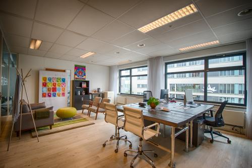 Kantoorruimte huren op Tolnasingel 1-3, Bodegraven. Gemeubileerd kantoor met grote ramen die veel licht binnenlaten, ingericht met bureau's en stoelen. Gezellige sfeer met planten en kunst aan de muur.