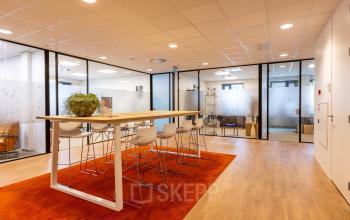 Moderne kantoorpand met glazen wanden en hoge houten tafels op een oranje tapijt. Ideale werkplek voor kantoorruimte huren in Bodegraven, Marktstraat 1-7.