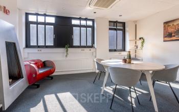 Lichte kantoorruimte met grote ramen, ronde tafel met stoelen en een decoratieve open haard in kantoorpand op Druivenstraat 25-31, Breda Oost. Ideaal voor kantoorruimte huren.