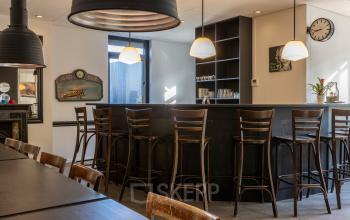 Een moderne kantoorruimte te huur in Breda Oost aan de Druivenstraat 25-31. Het interieur is voorzien van hoge houten stoelen aan een bar en een tafel. De ruimte is uitnodigend en functioneel.