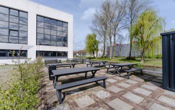Buitenzicht van kantoor op Rietbaan 2-12, Capelle aan den IJssel met een betegeld terras en houten picknicktafels. Kantoorruimte huren in groene omgeving.