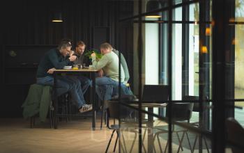 Zakelijke bijeenkomst in een moderne kantoorruimte op Rietbaan 2-12, Capelle aan den IJssel. Drie personen aan een hoge tafel in een gemeubileerde ruimte met veel lichtinval.