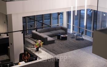 Moderne entree met zithoek in kantoorpand aan Rijnzathe 6, De Meern. Persoon bij de balie bezig met werk. Hoogwaardige inrichting, perfect voor wie kantoorruimte wil huren.